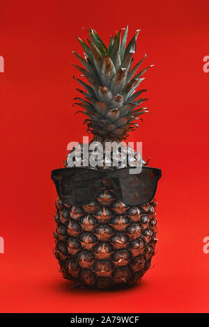 Funny ananas in occhiali da sole. Lay piatto su carta rossa sullo sfondo. ananas con gli occhiali su sfondo rosso. cool ananas Foto Stock