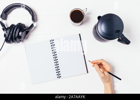 Vista superiore femmina scrittura a mano vuoto foglio notebook accanto al caffè e le cuffie sul tavolo bianco, laici piatta Foto Stock