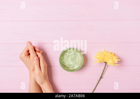 Donna con le mani in mano vicino a fresco fiore giallo e vaso di aloe gel cosmetico rosa sul tavolo di legno, piatto laici. Foto Stock