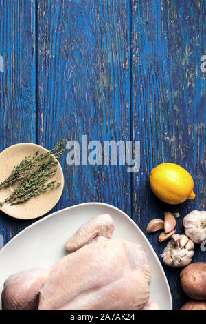 Il pollo crudo in una piastra bianca su sfondo blu sullo sfondo di legno con limone e un rametto di timo e vari ingredienti, laici piatta Foto Stock