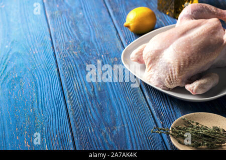 Il pollo crudo in una piastra bianca su sfondo blu sullo sfondo di legno con limone e un rametto di timo e vari ingredienti, vista laterale Foto Stock