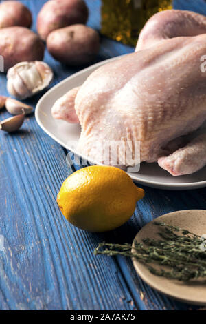 Il pollo crudo in una piastra bianca su sfondo blu sullo sfondo di legno con limone e un rametto di timo e vari ingredienti, vista laterale Foto Stock