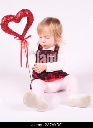 Piccola bionda toddler ragazza con grandi occhi blu in abito rosso gioca con la palla di Natale,seduta sul pavimento, sfondo bianco.ritratto, close-up isolato Foto Stock