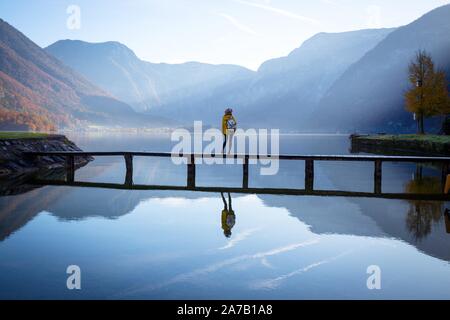 Ragazza turista in un cappello e con uno zaino sorge su un ponte di legno su un lago di montagna nelle prime ore del mattino. bellissimo paesaggio e di riflessione Foto Stock