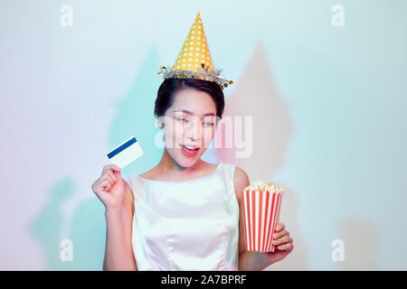 Ritratto di giovane contentissimo donna attraente in abito bianco guardare film, tenendo la benna di popcorn e una carta di credito isolato su bianco backgroun Foto Stock