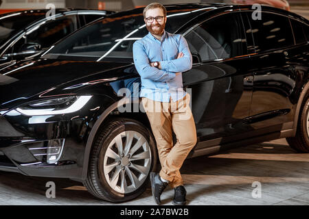 Corpo pieno ritratto di un felice giovane uomo in piedi vicino a una nuova auto elettrica presso lo showroom Foto Stock