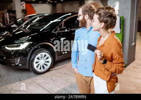 Ritratto di una coppia felice in piedi insieme con una chiave da una nuova auto elettrica presso lo showroom della concessionaria auto. Concetto di acquistare eco-friendly e auto moderne Foto Stock
