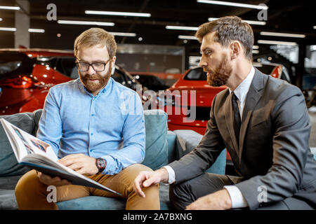 Uomo con sales manager scegliendo auto o auto con finiture in catalogo stampato sul comodo divano nella concessionaria auto Foto Stock