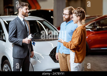 Sales Manager mostra auto stazione di ricarica per una coppia giovane, vendita di auto elettriche nello showroom. Concetto di acquistare eco-friendly auto per la famiglia Foto Stock