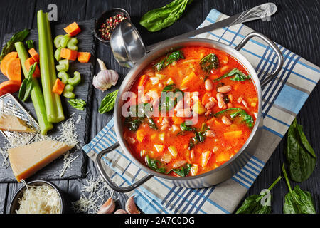 Fagioli borlotti zuppa con sedano, verdure, spinaci, il parmigiano e i pomodori in una pentola di metallo su un tavoli in legno nero, gli ingredienti su una pietra nera Foto Stock