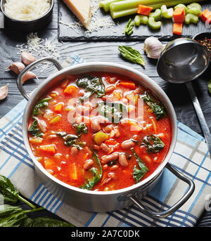 Toscana fagioli borlotti zuppa con sedano, verdure, spinaci, il parmigiano e i pomodori in una pentola di metallo su un tavoli in legno nero, gli ingredienti su un nero Foto Stock