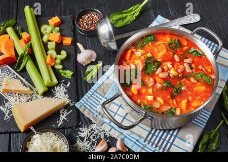 Italiano saporita minestra di fagioli di mirtillo palustre, sedano, verdure, spinaci, il parmigiano e i pomodori in una pentola di metallo su un tavoli in legno nero, ingredienti su Foto Stock