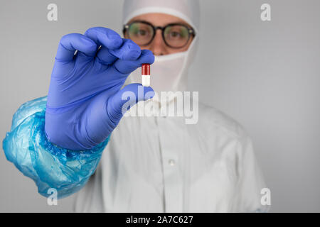 Giovane donna con gli occhiali che indossa bianca sterile indumenti di protezione e guanti blu tenendo un rosso e bianco pillola di fronte a lei Foto Stock