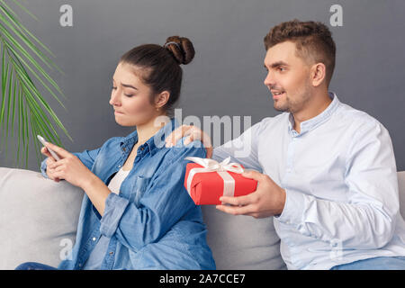 Coppia giovane uomo sorridente felice dando presente casella tocca lo spallamento della donna che gioca indifferente dello smartphone seduto sul divano studio isolato su grigio ba Foto Stock
