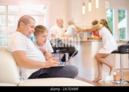 Il figlio e il padre sono sedute sul divano, guardando il laptop, gioiose emozioni da ciò che egli vedeva, in cucina la mamma con gli amici e una piccola Foto Stock