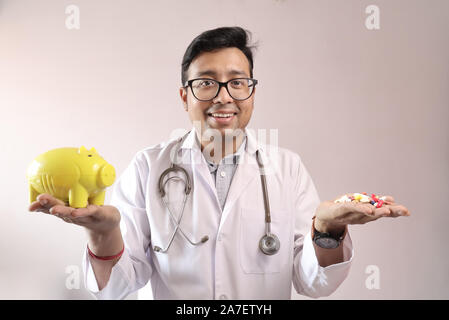 Indiano maschio medico in camice bianco e stetoscopio con un giallo salvadanaio e medicina pillole in mano. spese mediche Concetto di immagine Foto Stock