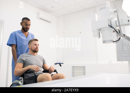 Giovane uomo con la gamba malata seduto su una sedia a rotelle nella parte anteriore di attrezzature mediche Foto Stock