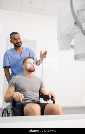 Dottore in uniforme blu che mostra il paziente su una sedia a rotelle nuove apparecchiature medicali Foto Stock