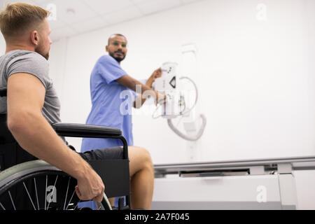 Giovane uomo con gamba rotta seduto su una sedia a rotelle e guardando il radiologo Foto Stock