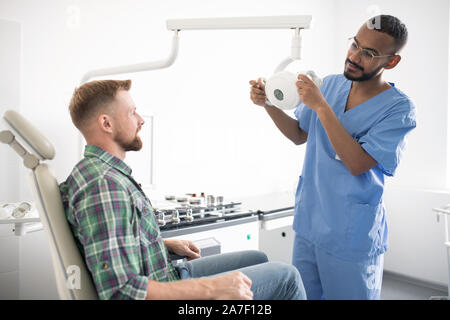 Giovane medico in uniforme di contenimento di apparecchiature mediche o lampada di fronte al paziente Foto Stock