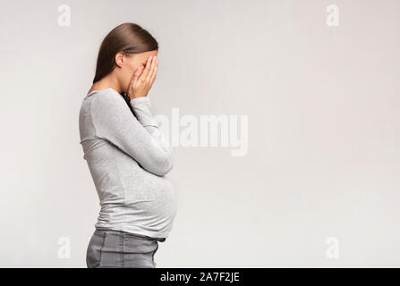 Premuto donna incinta che ricopre la faccia in piedi su sfondo grigio Foto Stock