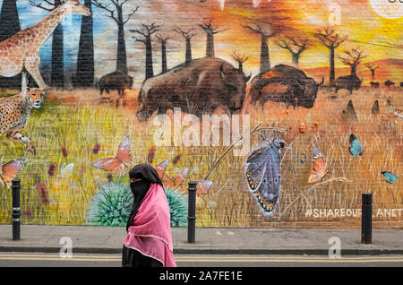 Un meraviglioso pezzo di arte di strada adorna una parete in Bethnal Green, East London REGNO UNITO. Passato a piedi è una donna nel tradizionale abito islamico. Foto Stock