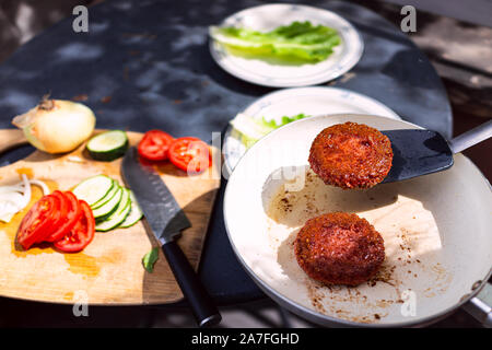 Servire due cotti a base di carne vegan polpette di salsiccia su ceramica nonstick padella con olio grasso pinneggiata spatola e i pomodori tagliati a fette di burger Foto Stock