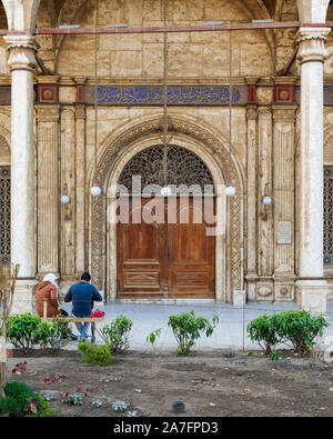 Il Cairo, Egitto - 10 Gennaio 2016: ornati in legno porta su marmo bianco parete decorata con coppia giovane seduto sulla panchina da giardino. entrata laterale di Muhammad Ali moschea, la Moschea di alabastro, Cittadella del Cairo Foto Stock