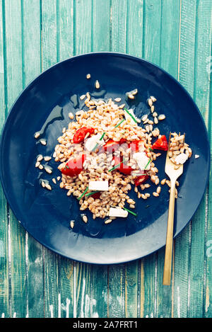 Insalata di farro fatta con scritto cotte ,pomodori ciliegia, erba cipollina e formaggio feta formaggio greco, la placcatura con una forcella dorata su una piastra di blu, , vista dall'alto su un bianco Foto Stock