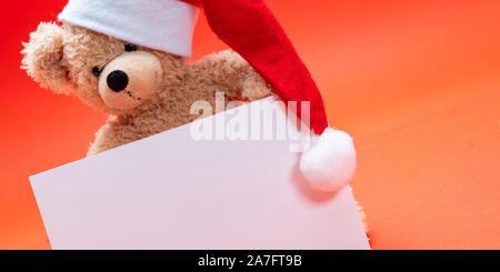 Scheda di natale template. Orsacchiotto di peluche con santa hat tenendo un foglio bianco, rosso colore di sfondo, spazio copia Foto Stock