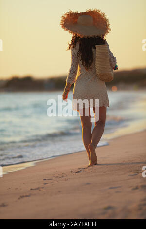 A piena lunghezza ritratto della moderna medioevo donna in abito bianco e il cappello di paglia sulla spiaggia al tramonto a piedi. Foto Stock