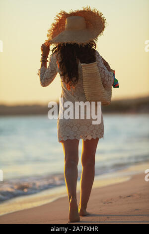 A piena lunghezza Ritratto di giovane donna in abito bianco e il cappello di paglia sul Seacoast al tramonto a piedi. Foto Stock