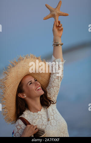 Sorridente moderno 40 anno vecchia donna in abito bianco e il cappello di paglia sulla spiaggia al tramonto holding starfish. Foto Stock