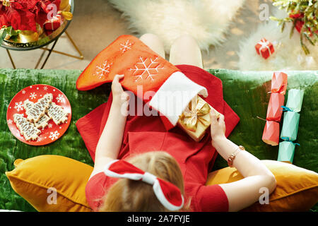 Stagione di vacanze. Vista superiore della elegante piccola principessa in abito rosso seduto sul divano nel soggiorno moderno a Natale prendendo un regalo di Natale b Foto Stock