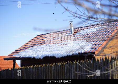 La prima neve sul tetto di tegole. Gli uccelli sono volare nel cielo blu. Log in legno recinzione. Foto Stock