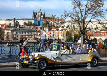 I turisti di Praga in auto d'epoca taxi auto finte veterane corsa persone giro turistico Smetanovo nabrezi che si affaccia sul paesaggio urbano del castello di Praga sopra il fiume Moldava Foto Stock