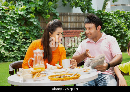 La famiglia felice i genitori leggendo il giornale e parlando con il figlio avente la prima colazione nel cortile di casa loro Foto Stock