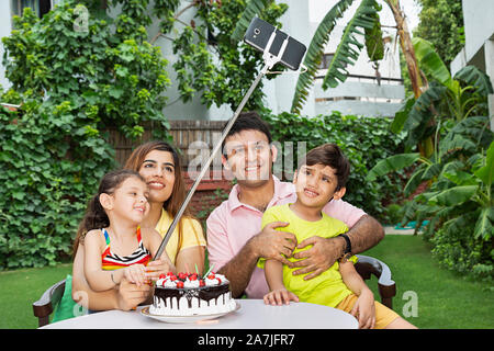 La famiglia felice tenendo selfie foto da smartphone con selfie-stick durante il celebrare la festa di compleanno nel cortile di casa loro Foto Stock