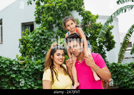 Felice famiglia giovane madre e padre portando la sua figlia di capretto sulle spalle mentre godendo in cortile vicino a casa loro Foto Stock