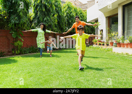 Felice famiglia indiana i genitori di due bambini con Arms-Outstretched correndo insieme In-Garden vicino a casa loro Foto Stock