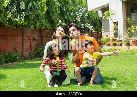 Felice Famiglia giovani genitori e due bambini soffiaggio in bolla wand facendo sapone palloncini in-cortile vicino a loro casa Foto Stock