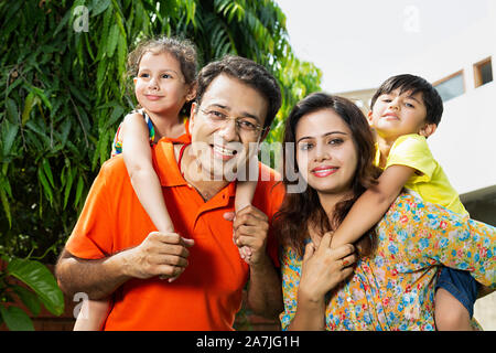 Felice Indian giovane famiglia i genitori dando piggyback ride per bambini nel cortile di casa loro Foto Stock