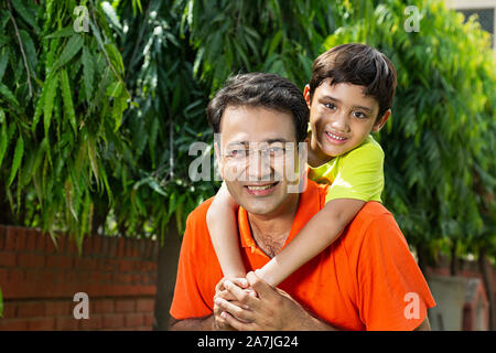 Famiglia indiana sorridente padre dando per il suo nero piccolo figlio di piggyback ride presso il giardino esterno Foto Stock