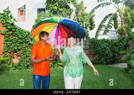 Felice Coppia di anziani con ombrellone in piedi sotto la pioggia e parlando godendo la pioggia nel giardino di casa Foto Stock