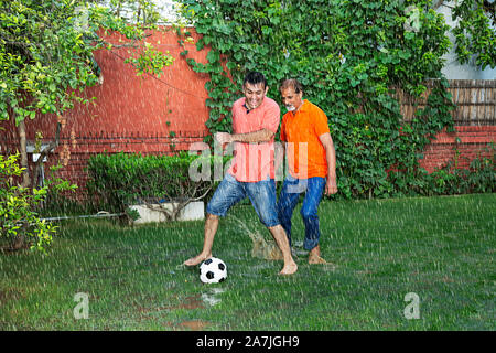 Felice famiglia indiana padre senior e adulti-figlio che giocano a calcio palla in gioco-la pioggia nel giardino di casa Foto Stock