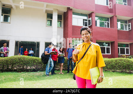 Femmina giovane studente di college Azienda Libro mentre mostra Thumbs up e gli studenti In-The-At-Campus sfondo Foto Stock