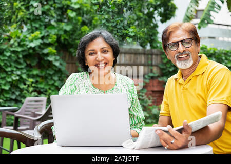 Felice vecchia coppia maschio e femmina utilizzando laptop e leggere il giornale in-cortile di casa loro Foto Stock