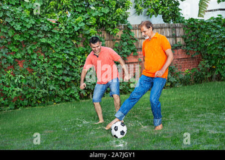 Adult-Son e invecchiato padre giocando a calcio In-The pioggia Of-Their In-Park House Foto Stock