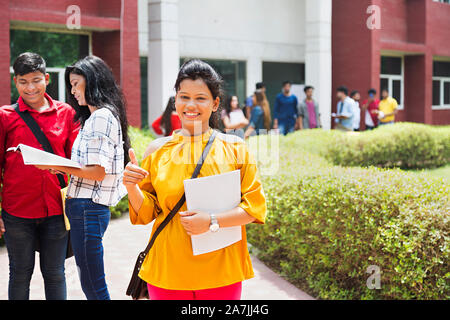 Femmina giovane studente di college Holding-Book mostrando pollice in su con gli studenti amici In-Outside Campus Foto Stock