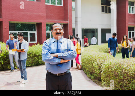 Uomo Senior College insegnante permanente Arms-Crossed con Students-In-The-Background In-Outside Campus Foto Stock